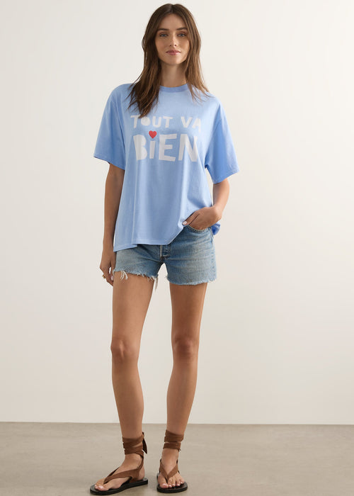 A woman wearing a light blue oversized graphic t-shirt with the French phrase 'TOUT VA BIEN' printed in white block letters with a small red heart accent, paired with blue denim shorts.