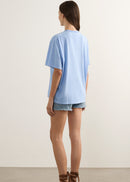 A woman shown from the back wearing an oversized light blue short-sleeve t-shirt, denim shorts, and brown strappy sandals, standing against a plain off-white wall.