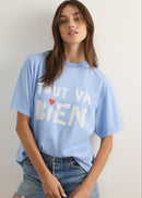 A woman wearing a light blue oversized graphic t-shirt with the French phrase 'TOUT VA BIEN' printed in white block letters with a small red heart accent, paired with blue denim shorts.