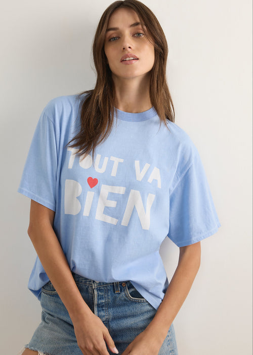 A woman wearing a light blue oversized graphic t-shirt with the French phrase 'TOUT VA BIEN' printed in white block letters with a small red heart accent, paired with blue denim shorts.