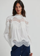 A woman wearing a white long-sleeve blouse with intricate lace detailing at the mock neck, shoulders, and hem, paired with dark blue jeans, against a light grey studio background.