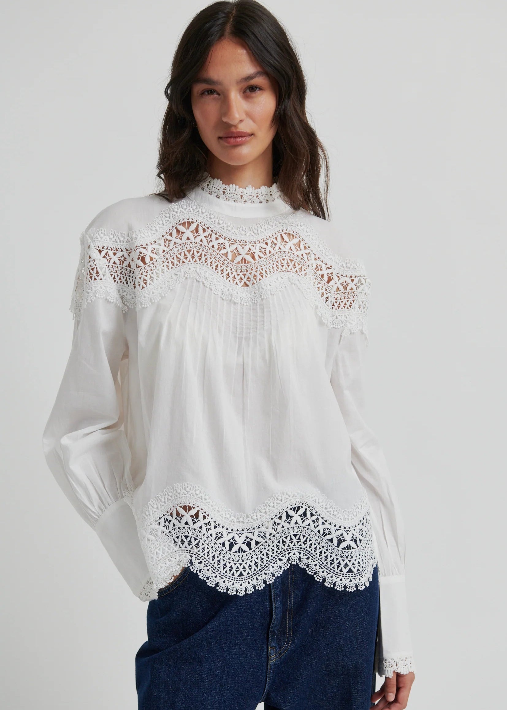 A woman wearing a white long-sleeve blouse with intricate lace detailing at the mock neck, shoulders, and hem, paired with dark blue jeans, against a light grey studio background.