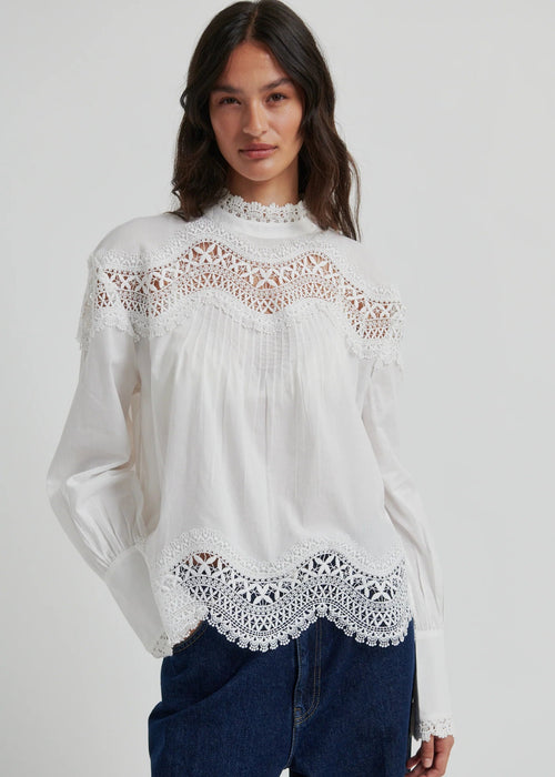 A woman wearing a white long-sleeve blouse with intricate lace detailing at the mock neck, shoulders, and hem, paired with dark blue jeans, against a light grey studio background.