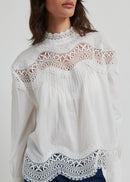 A woman wearing a white long-sleeve blouse with intricate lace detailing at the mock neck, shoulders, and hem, paired with dark blue jeans, against a light grey studio background.