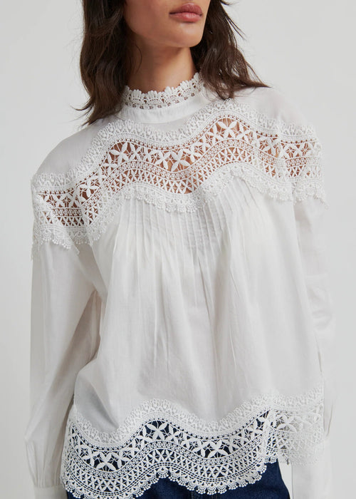 A woman wearing a white long-sleeve blouse with intricate lace detailing at the mock neck, shoulders, and hem, paired with dark blue jeans, against a light grey studio background.