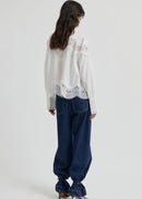A woman wearing a white long-sleeve blouse with intricate lace detailing at the mock neck, shoulders, and hem, paired with dark blue jeans, against a light grey studio background.