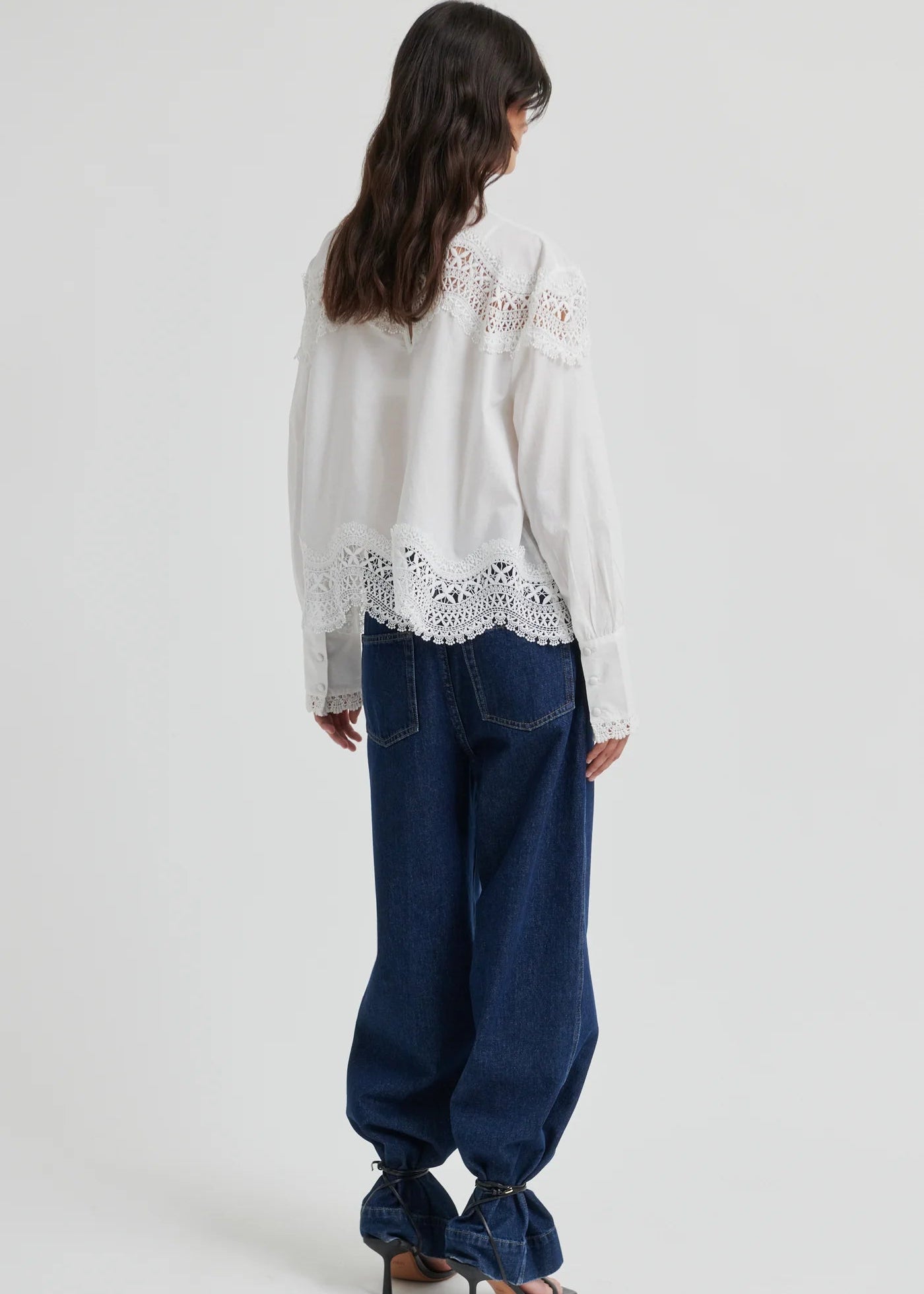 A woman wearing a white long-sleeve blouse with intricate lace detailing at the mock neck, shoulders, and hem, paired with dark blue jeans, against a light grey studio background.