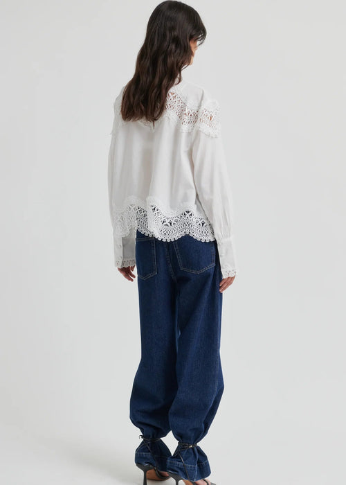 A woman wearing a white long-sleeve blouse with intricate lace detailing at the mock neck, shoulders, and hem, paired with dark blue jeans, against a light grey studio background.