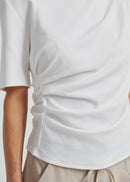 A woman modelling a white short-sleeve top with a draped, asymmetrical neckline and gathered front detail, paired with light beige drawstring pants, against a plain light grey studio background.