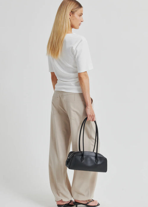 A woman modelling a white short-sleeve top with a draped, asymmetrical neckline and gathered front detail, paired with light beige drawstring pants, against a plain light grey studio background.