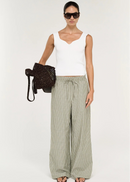 A model wearing a white ribbed knit scoop-neck tank top and green and white vertically striped trousers, accessorized with black sunglasses and a black woven shoulder bag, against a plain off-white background.