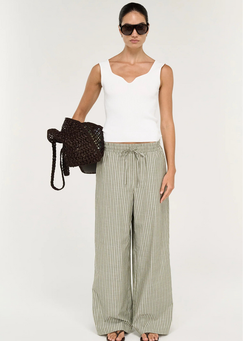 A model wearing a white ribbed knit scoop-neck tank top and green and white vertically striped trousers, accessorized with black sunglasses and a black woven shoulder bag, against a plain off-white background.