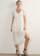 A woman modelling a white short-sleeve midi dress with lace detailing at the neckline and hem, paired with black strappy flat sandals, standing against a plain white wall.