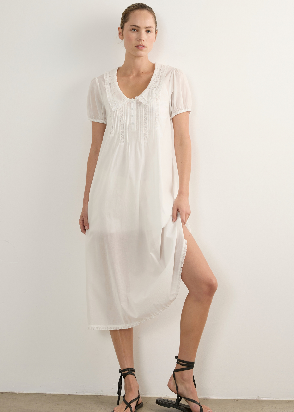 A woman modelling a white short-sleeve midi dress with lace detailing at the neckline and hem, paired with black strappy flat sandals, standing against a plain white wall.