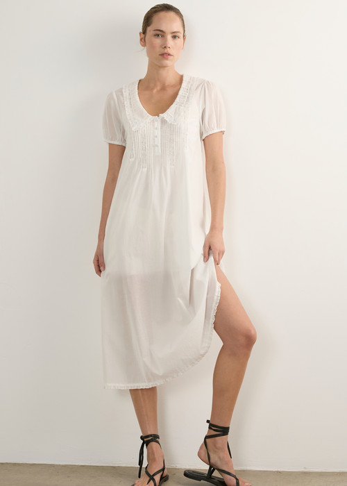 A woman modelling a white short-sleeve midi dress with lace detailing at the neckline and hem, paired with black strappy flat sandals, standing against a plain white wall.