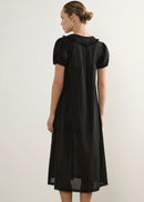 A woman modelling a black short-sleeve midi dress with a ruffled Peter Pan collar and gathered front, standing against a plain light grey studio background.