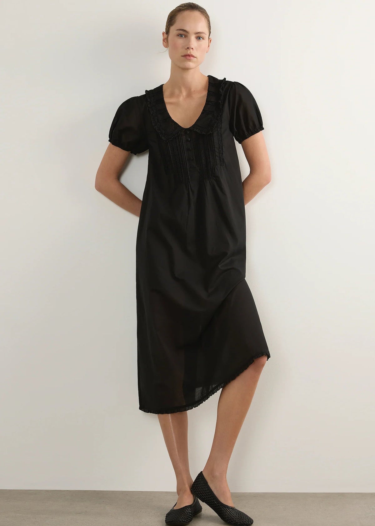 A woman modelling a black short-sleeve midi dress with a ruffled Peter Pan collar and gathered front, standing against a plain light grey studio background.