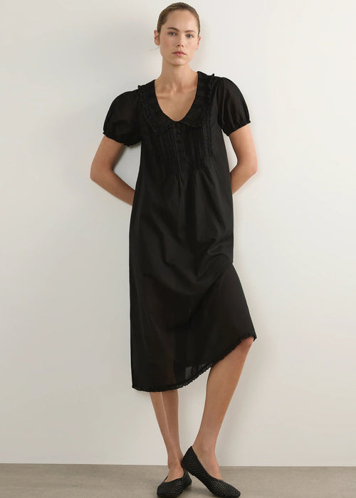 A woman modelling a black short-sleeve midi dress with a ruffled Peter Pan collar and gathered front, standing against a plain light grey studio background.