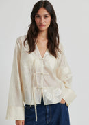 Woman wearing a light-coloured blouse with long sleeves and a tie detail on a white background