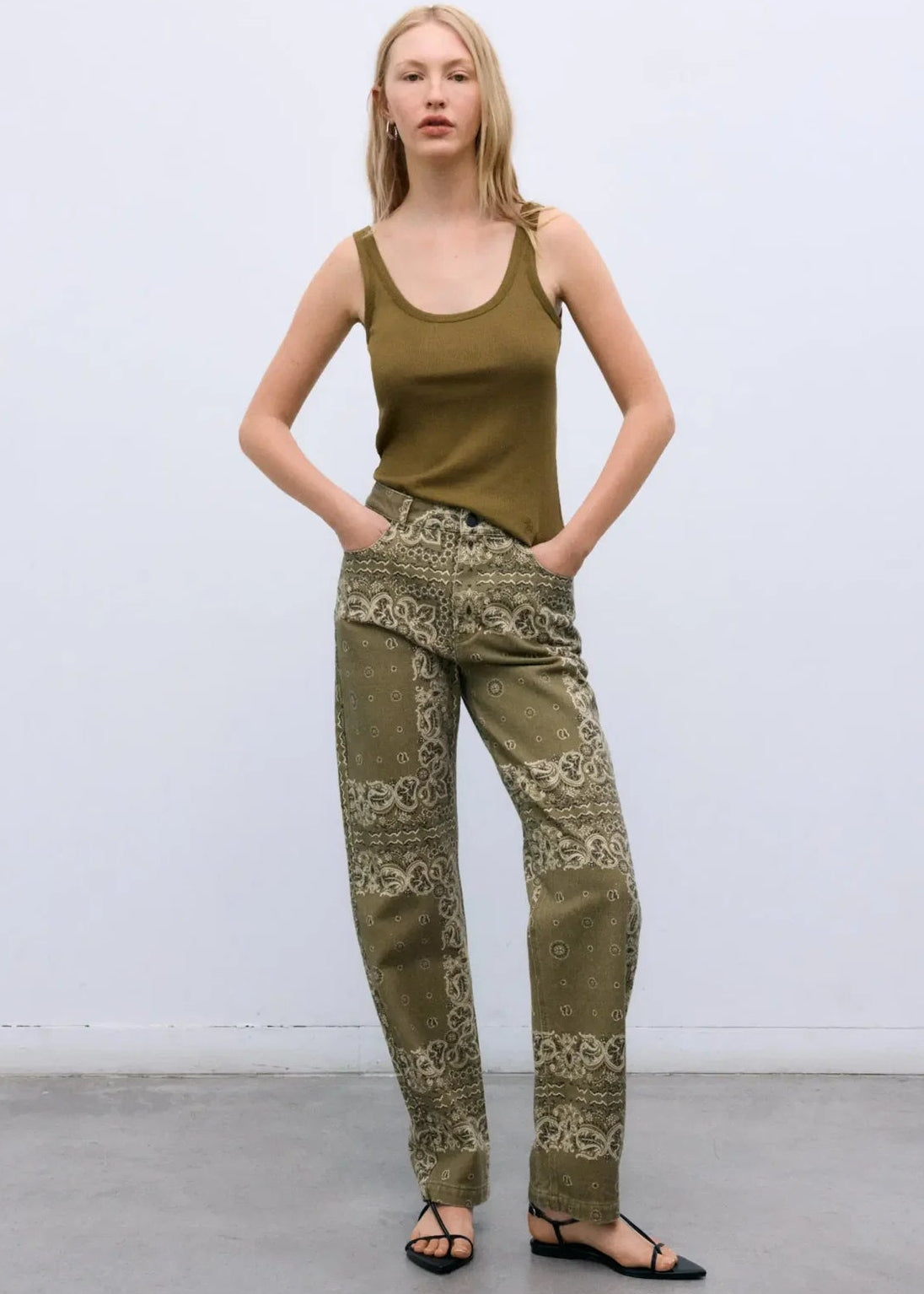 A woman stands against a light gray studio wall wearing olive green patterned pants with a matching olive green tank top and black flat sandals.