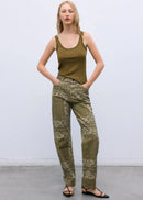 A woman stands against a light gray studio wall wearing olive green patterned pants with a matching olive green tank top and black flat sandals.