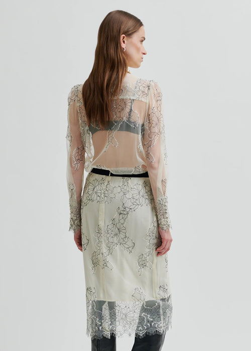 Model wearing a sheer blouse with floral embroidery against a white background