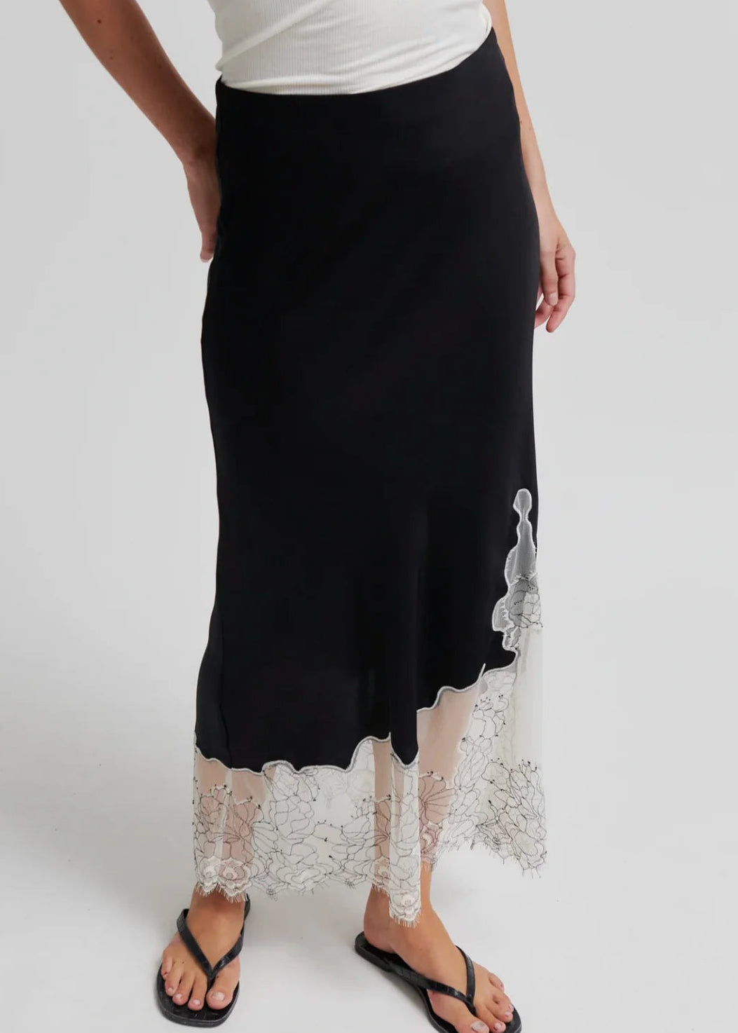 A black midi skirt with a sheer white lace hem and floral applique, worn by a model with a white top and black flip-flops against a light grey studio background.