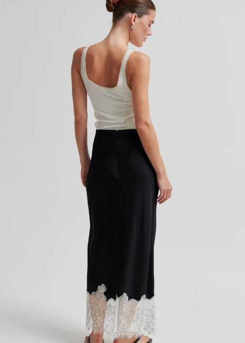 A woman shown from the back wearing a sleeveless white top and a long black skirt with a white lace hem, photographed against a light grey studio background.