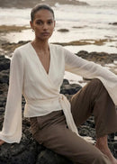 A model sits on dark volcanic rocks by the ocean, wearing a cream wrap-style long-sleeve top with a tie waist and brown wide-leg trousers.