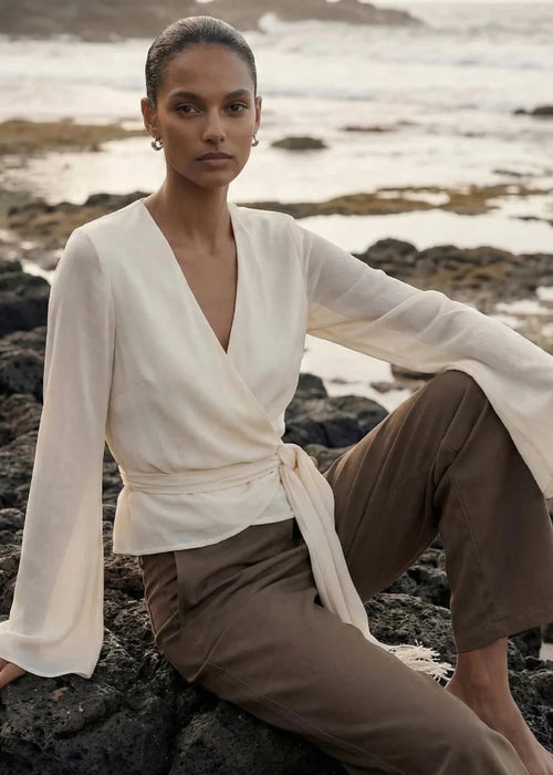 A model sits on dark volcanic rocks by the ocean, wearing a cream wrap-style long-sleeve top with a tie waist and brown wide-leg trousers.