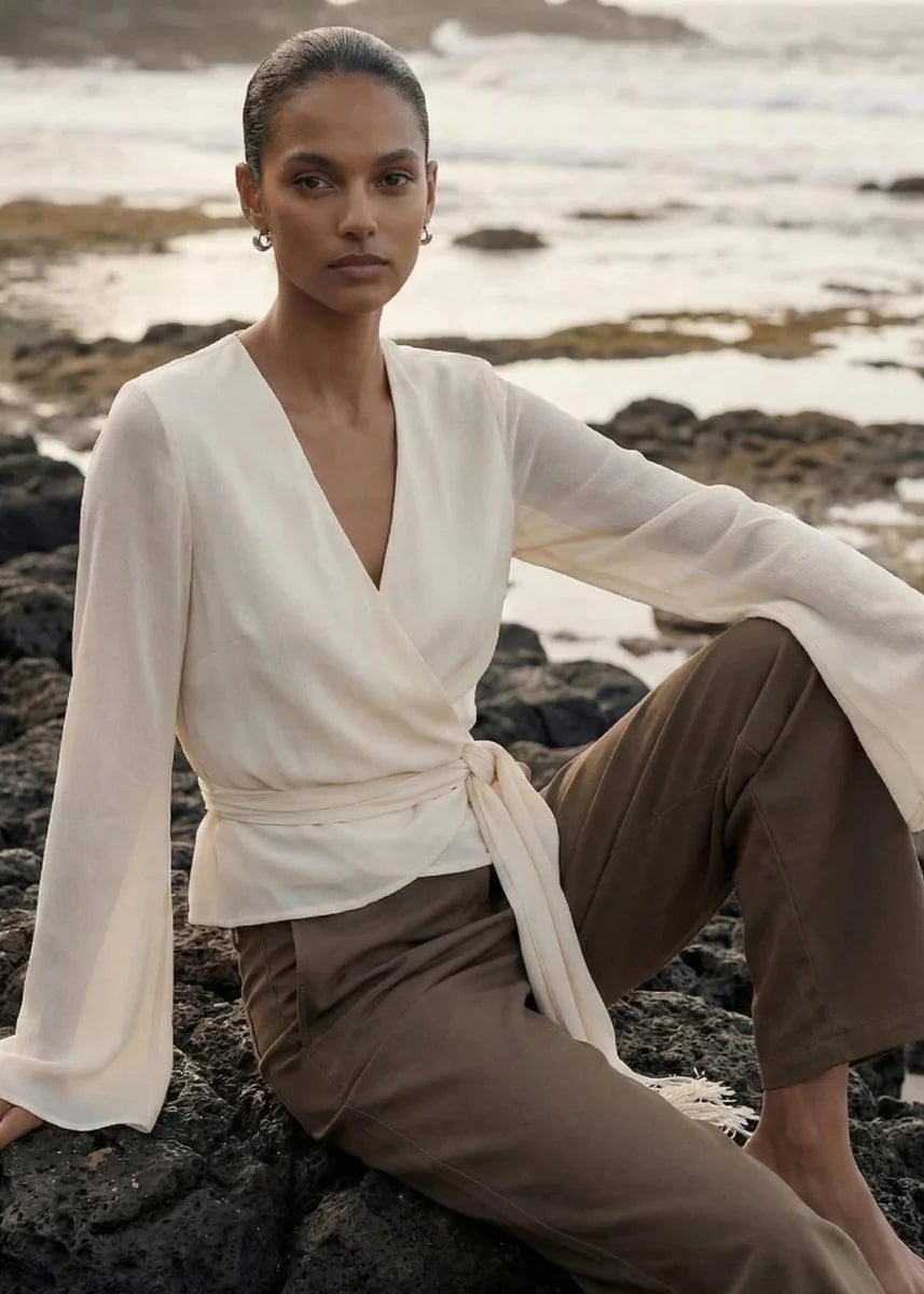A model sits on dark volcanic rocks by the ocean, wearing a cream wrap-style long-sleeve top with a tie waist and brown wide-leg trousers.