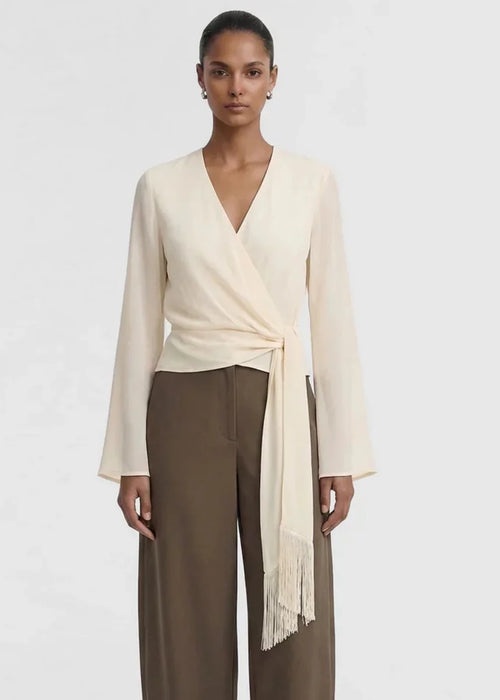 A model wearing a cream-colored wrap top with long sleeves and a fringed tie detail, paired with brown wide-leg trousers, against a plain light grey background.