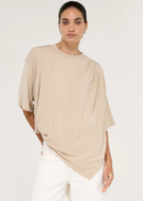 A model stands in profile wearing a loose, draped beige short-sleeve top, white shorts, and dark brown flip-flops against a plain light grey studio background.