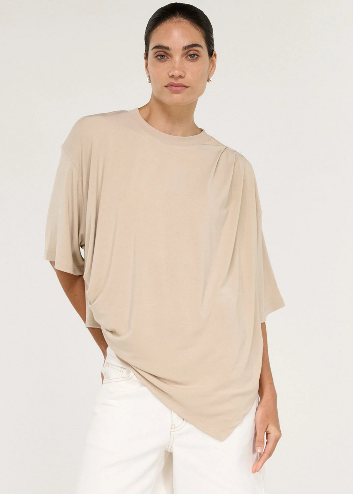 A model stands in profile wearing a loose, draped beige short-sleeve top, white shorts, and dark brown flip-flops against a plain light grey studio background.