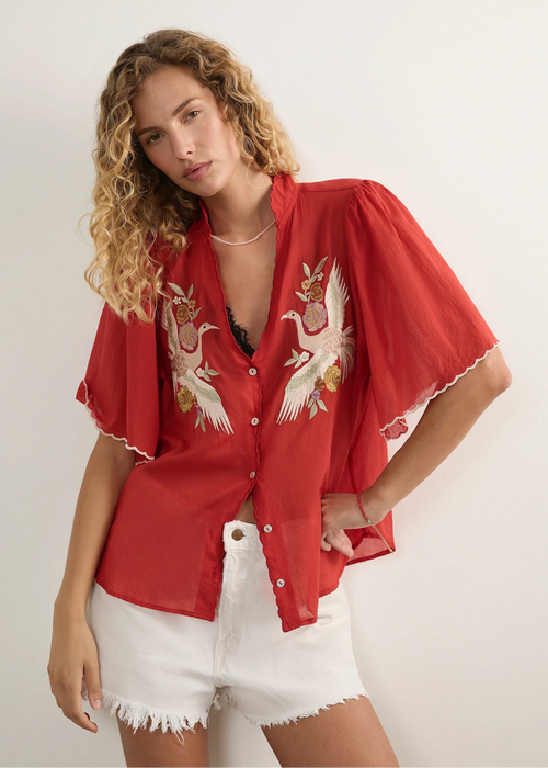 A woman modelling a red short-sleeve button-up shirt with white and gold crane embroidery on the front, paired with white denim shorts against a plain light background.